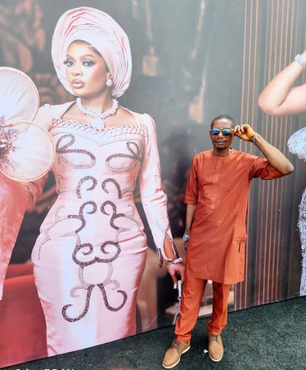 Emmanuel Obasi poses with a photo of May Yul-Edochie in the background.